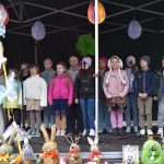 Dzieci w kolorowych kurtkach śpiewają na scenie. W tle ciemne zadaszenie z wiszącymi kwiatami. Z przodu dekoracje z zajączkami.