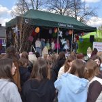 Grupa ludzi stoi przed sceną z zielonym daszkiem z napisem Gmina Dobre. Na scenie występują dzieci. W tle widoczne są chmury na niebieskim niebie.