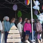 Grupa dzieci w kolorowych kurtkach śpiewa na scenie wiosennego festynu. Tło zdobią duże, jasne kwiaty i ozdoby. Scena jest pod zielonym namiotem.