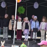 Grupa dzieci śpiewa na scenie pod zadaszeniem, ubrana w zimowe kurtki, szaliki i czapki. W tle kolorowe dekoracje, jak duże, papierowe kwiaty. Na pierwszym planie widoczne są rękodzielnicze ozdoby z motywem z