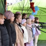 Grupa dzieci śpiewa do mikrofonów na scenie. Ubrane są ciepło, tło to zielona płachta z dekoracjami. Dzieci stoją obok siebie, skupione na występie.