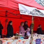 Na zdjęciu widoczny jest czerwony namiot z banerem KGW Małomiasteczkowi. Przed namiotem stoi stół z rękodziełem, przysmakami i dekoracjami. W tle kilka osób rozmawia, a dziecko w jasnoniebieskiej kurtce