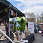 Scena z kolorowymi dekoracjami i dziećmi śpiewającymi w chórze. Na pierwszym planie figurki królików z wikliny. W tle targowisko i drzewa.