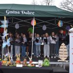 Grupa dzieci śpiewa na scenie pod zielonym namiotem z napisem Gmina Dobre. Na pierwszym planie dekoracje wielkanocne. Obok stoi banner Mazowsze, a w tle widać kolorowe lampiony.