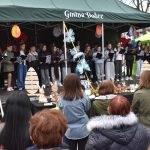 Grupa młodzieży na scenie w zielonym namiocie, śpiewa z nutami. Przed nimi ozdoby wielkanocne, a wokół stoją widzowie. Scena ozdobiona kolorowymi lampionami.