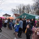 Na zdjęciu jest grupa ludzi oglądająca wydarzenie na świeżym powietrzu. W tle zielony namiot i kolorowe stoiska. Dookoła drzewa bez liści.