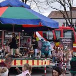 Kolorowa karuzela z dziećmi na tle czerwonego wozu strażackiego. Parasolka w kolorach niebieskim, zielonym i czerwonym. Wokół stoją dorośli, jest chłodny dzień.