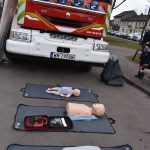 Czerwony wóz strażacki stoi na tle budynków; przed nim leżą manekiny do nauki resuscytacji na szarych matach.