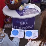 Dwie osoby trzymają ulotki na tle okrągłego stołu z napisem GRA TERENOWA. Na blacie leży koszyk z kolorowymi dekoracjami. Tło jest neutralne, z czerwonymi i fioletowymi akcentami.