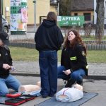 Dwóch strażaków na chodniku prowadzi pokaz pierwszej pomocy z użyciem manekinów. Na trawie w tle widoczna jest apteka. Ubrani są w czarne ubrania z oznaczeniami straży.