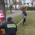 Dzieci trenują gaszenie pożaru z pomocą strażaka. Zimowe ubrania, szary dzień, zielone tło parku z drzewami i budynki.