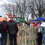 Ludzie oglądają wiosenny kiermasz na tle namiotów w różnych kolorach: pomarańczowym, zielonym i niebieskim. Drzewa bez liści stanowią tło. Drewniany płotek otacza centralne stoisko.