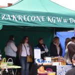 Stoisko z zielonym namiotem na targu, z różnokolorowymi koszykami na stole, przy którym stoi grupa osób. Za nimi napis ZAKRĘCONE KGW w Dobrem na tle. Obok widoczny szyld z napisem GRA TERENOWA.