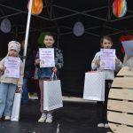 Trójka dzieci z dyplomami i torbami upominkowymi stoi na scenie pod ciemnym zadaszeniem. W tle kolorowe papierowe dekoracje. Na prawo kobieta w jasnym płaszczu trzyma kartkę.