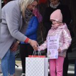 Osoba wręcza dyplom i prezent dziecku w różowym ubraniu. Tło ciemne, z ozdobami. Dziecko trzyma dyplom w rękach.