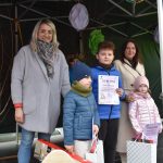 Grupa ludzi stoi pod zielonym namiotem. Dzieci trzymają dyplomy i torby z prezentami. Tło zdobią wiszące ozdoby.