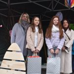 Cztery osoby pozują pod namiotem z kolorowymi dekoracjami. Dwie dziewczynki trzymają szare torby na nagrody. Klimat wydarzenia jest świąteczny.