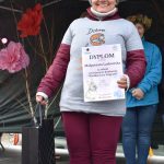 Osoba w szarej koszulce i bordowych spodniach trzyma dyplom i czarną torbę na tle dekoracji. Jest uśmiechnięta, z zimową czapką na głowie. Tło to ciemny namiot i kolorowe kwiaty papierowe.