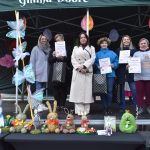 Grupa sześciu osób stoi na scenie pod zielonym namiotem z dyplomami. Przed nimi kolorowe, ręcznie robione zajączki wielkanocne. Tło zdobią kolorowe kwiaty i dekoracje.