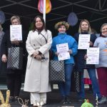 Na scenie stoi pięć kobiet z dyplomami i torbami na tle kolorowych dekoracji. Tło jest ciemne z wiszącymi ozdobami. Kobiety są ubrane w różnorodne stroje, w tym płaszcze i koszulki.