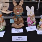 Na zdjęciu widać ręcznie robione zające wielkanocne ustawione rzędem na czarnym tle. każdy z nich jest ozdobiony różnymi materiałami, a przed nimi stoją kartki z imionami laureatów. Tło jest ciemne, co podkreśla kolorowe detale