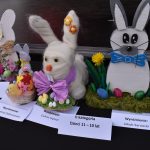 Na zdjęciu są trzy dekoracyjne zające wielkanocne z różnych materiałów. Każdy zając ma unikalne ozdoby, jak kolorowe jajka i kwiaty. W tle ciemne tło, a zające są wyróżnione certyfikatami.