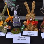 Cztery ręcznie wykonane zające z siana i wełny w różnych kolorach. Na czarnym tle widoczne kolorowe wstążki. Tabliczki z nagrodami dla dziecięcej kategorii wiekowej.