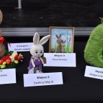 Trzy ręcznie wykonane zające w różnych stylach na tle czarnego stołu. Pierwszy jest brązowy, drugi biały, trzeci zielony. Obok zające stoją kartki z nagrodami dla twórców.