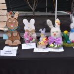 Na zdjęciu widać ręcznie wykonane dekoracje zajączków wielkanocnych w różnych kolorach. Stoją one na czarnym stole, a każda ma swoją tabliczkę z miejscem lub wyróżnieniem. W tle jest ciemne tło sceniczne, co skupia uwagę na dekor