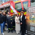 Grupa osób stoi przed czerwonym wozem strażackim z otwartymi skrytkami. Obok widać banner Ochotniczej Straży Pożarnej w Mlecinie. Tło stanowią drzewa i niebo.