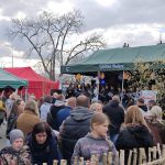 Na zdjęciu tłum ludzi zgromadzony przed sceną z zielonym namiotem. W tle są czerwone i zielone namioty, a niebo pokryte chmurami. Na scenie występują dzieci, a wokół znajdują się kwitnące gałązki.