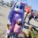 Na zdjęciu postać królika w fioletowym stroju trzyma kosz z jajkami, obok stoi dziecko w różowej kurtce. Tło to plac z grupą ludzi i czerwonym namiotem. Na pierwszym planie kwiaty i trawy ozdobne.