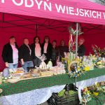 Grupa osób stoi pod różowym namiotem z napisem. Stół ozdobiony zielonym obrusem z czerwonymi wzorami, na którym znajdują się rękodzieła i jedzenie. W tle widać drzewa bez liści.