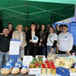 Grupa ludzi stoi pod zielonym namiotem. Przed nimi stół z przetworami i wypiekami. Tło to niebieskie i zielone płótno.