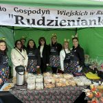 Sześć kobiet stoi przed zielonym namiotem z banerem Koło Gospodyń Wiejskich w Rudzience 'Rudzienianki'. Mają fartuchy z kwiatowym wzorem i opaski z dekoracjami. Na stole przed nimi znajdują się ciasta w plastikowych pude