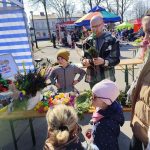Na zdjęciu widać grupę osób oglądających kolorowe, wielkanocne palmy na straganie z białymi i niebieskimi pasami. W tle są stoiska z jedzeniem i kolorowe parasole, a całość odbywa się na otwartym powietrzu. Pogoda jest sł