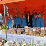 Grupa ludzi w niebieskich ubraniach stoi pod pomarańczowym namiotem. Na stole są ozdoby, wypieki i kwiaty. Tło to teren na zewnątrz z pojazdem.