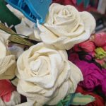 Bukiet z papieru składa się z białych róż i kolorowych kwiatów, otoczony zielonymi liśćmi. Widoczny niebieski klips. Tło jest rozmyte, z książkami.