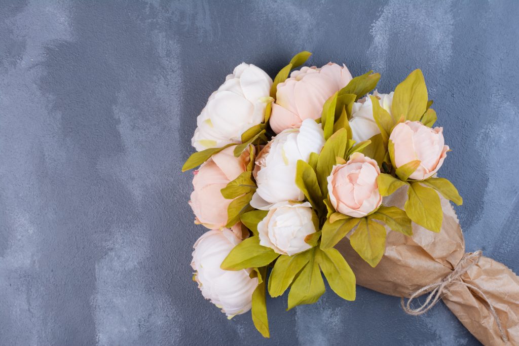 Bukiet jasnoróżowych i białych kwiatów owinięty w brązowy papier na szarej powierzchni. Liście w odcieniu zielonym. Tło matowe.