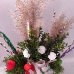 Bukiet wielkanocny z zielonymi gałązkami i białymi kwiatami na jasnym tle. Dodatki to czerwone wstążki i ozdobne suszone trawy. Kolory stonowane, zielono-beżowo-czerwone.