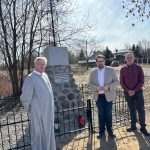 1000052521 Trzech mężczyzn stoi przy kamiennym pomniku z metalowym krzyżem. Tło to bezlistne drzewa i błękitne niebo. Pomnik jest ogrodzony czarnym płotem.