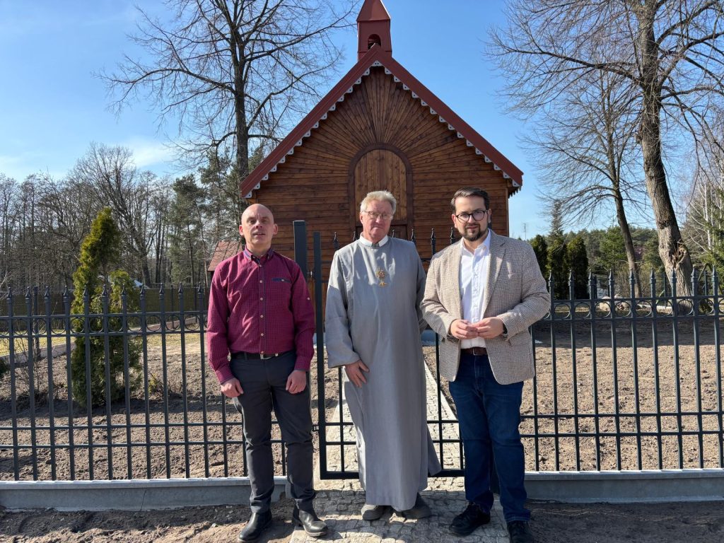 Trzech mężczyzn stoi przed drewnianą kaplicą na tle ogrodzenia. Jeden mężczyzna ma szarą sutannę, a dwóch nosi eleganckie ubrania. W tle widać drzewa i czyste niebo.