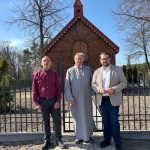 1000052522 Trzech mężczyzn stoi przed drewnianą kaplicą na tle ogrodzenia. Jeden mężczyzna ma szarą sutannę, a dwóch nosi eleganckie ubrania. W tle widać drzewa i czyste niebo.