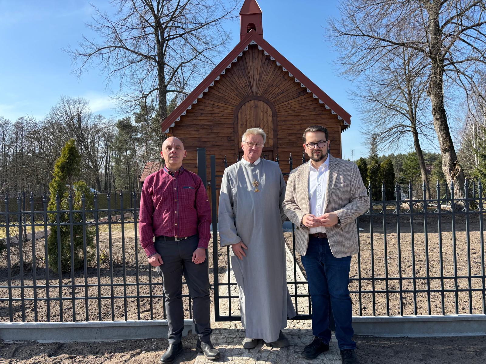Trzech mężczyzn stoi przed drewnianą kaplicą na tle ogrodzenia. Jeden mężczyzna ma szarą sutannę, a dwóch nosi eleganckie ubrania. W tle widać drzewa i czyste niebo.