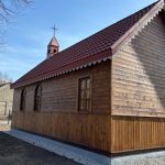 1000052523 Drewniany kościół z czerwonym dachem na tle nieba. Ma okna w ostrołukowym stylu. Obok widać budynek i ogrodzenie.