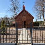 1000052525 Drewniany kościółek na tle błękitnego nieba i drzew. Czarne ogrodzenie i brukowana ścieżka prowadzą do wejścia. Otoczenie jest zadbane i spokojne.