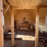 1000052527 Wnętrze drewnianej kaplicy z jasnym, naturalnym wykończeniem. Na środku znajduje się ołtarz, otoczony balustradą. Po prawej stronie widać ławki i duże okno wpuszczające światło.