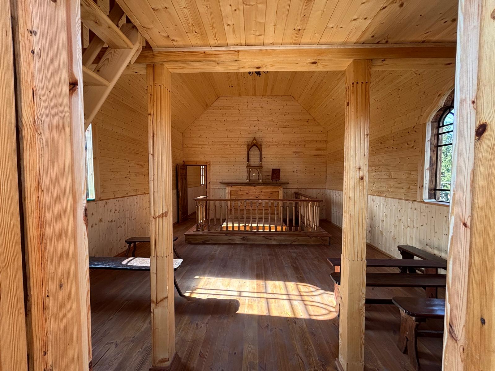 1000052527 Wnętrze drewnianej kaplicy z jasnym, naturalnym wykończeniem. Na środku znajduje się ołtarz, otoczony balustradą. Po prawej stronie widać ławki i duże okno wpuszczające światło.