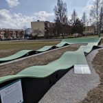20260319_115447 Skatepark z zielonymi falistymi rampami na tle bloków i drzew. Niebo zachmurzone, teren otoczony żwirem. Tablica informacyjna z przodu.
