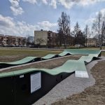 20260319_115454 Tor rowerowy z zielonymi falistymi rampami na tle bloków i drzew. Niebo jest lekko pochmurne. Teren jest trawiasty.